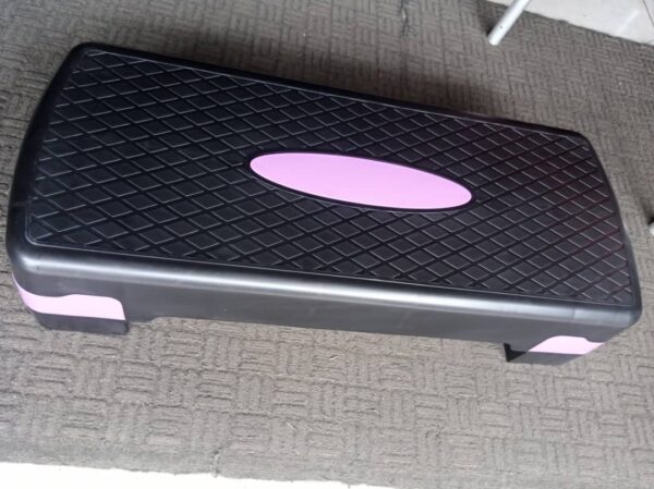 A black aerobic step board with a pink oval in the center. The board has a textured surface and raised edges for stability.