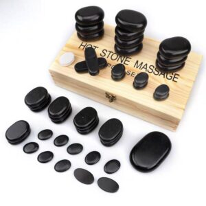 A wooden box filled with black, smooth stones for hot stone massage. The stones are arranged in stacks and are accompanied by a white candle and a small wooden tray. The label "HOT STONE MASSAGE" is printed on the box.