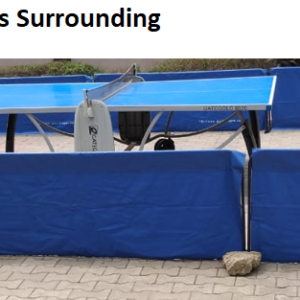 A blue and white table tennis table surrounded by blue barriers. There is a stone on the ground near one of the barriers.