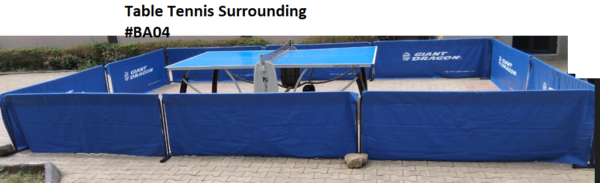 A blue and white table tennis table surrounded by blue barriers. There is a stone on the ground near one of the barriers.